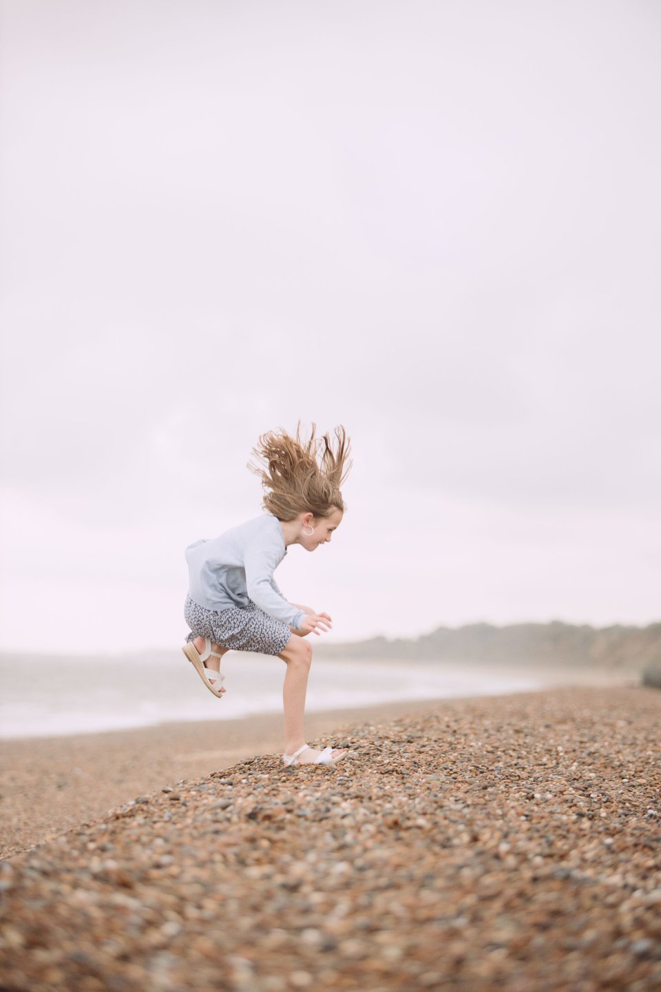 A person jumping on a pebbly beach with hair flying upward against an overcast sky.