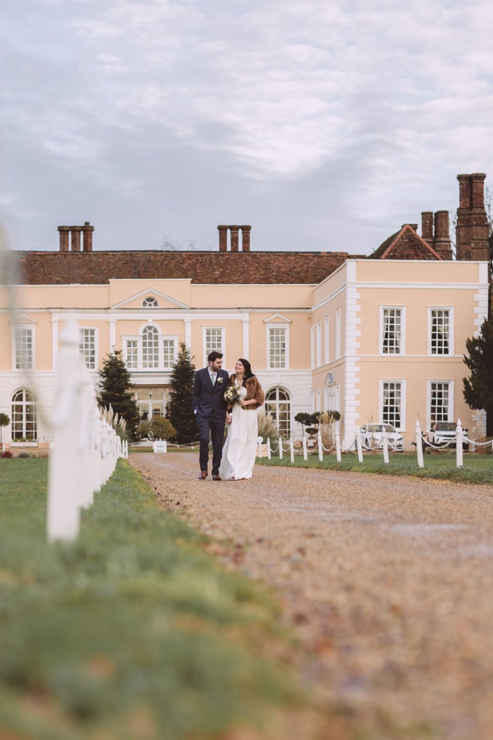 A bride and groom walk hand in hand away from Suffolk Wedding venue Hintlesham Hall with the hall in the background