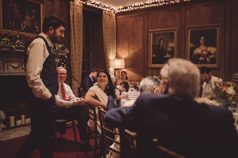A bride laughs at her husband who stands over her at her wedding breakfast at Hintlesham Hall