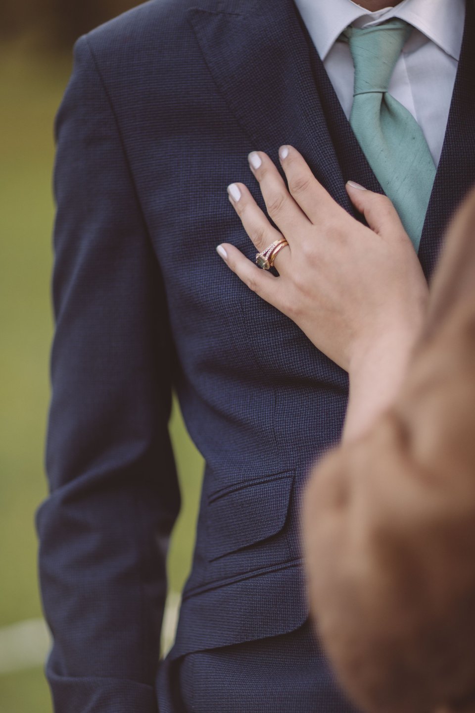 A close-up photograph of a bride's hand placed on her groom's chest with her rings visible.
