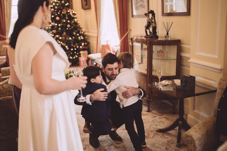 A groom crouching down as he embraces two young boys whilst his bride looks on in Hintlesham Hall drawing room