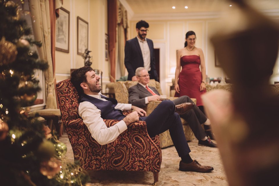 A groom laughs at his wedding witting in an armchair in a drawing room at Hintlesham Hall.
