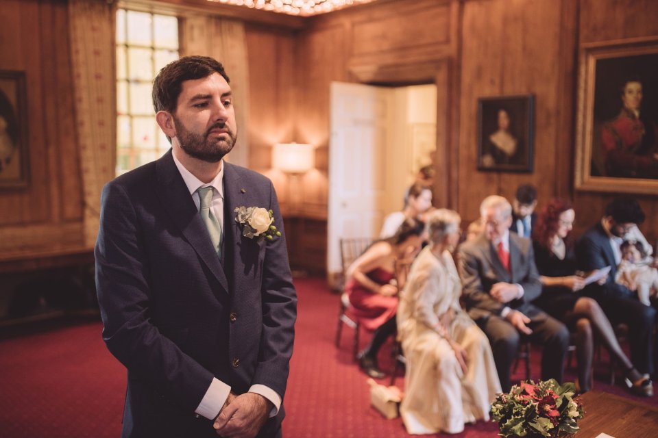 A groom stands at the front of the ceremony room in Hintlesham Hall awaiting the arrival of his bride at his wedding