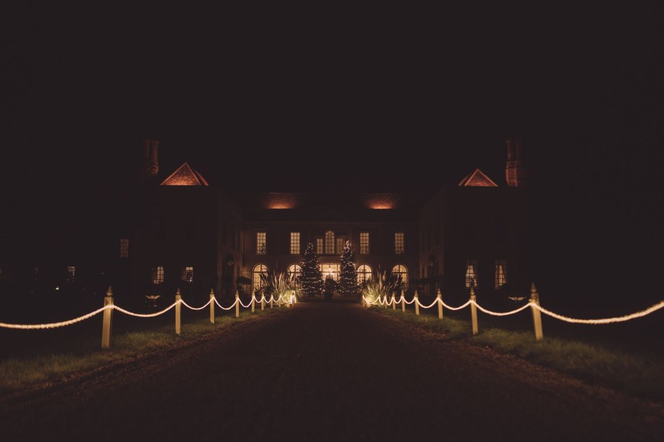 A night time exterior photograph of Hintlesham Hall in Suffolk with lit windows and chains lining the driveway