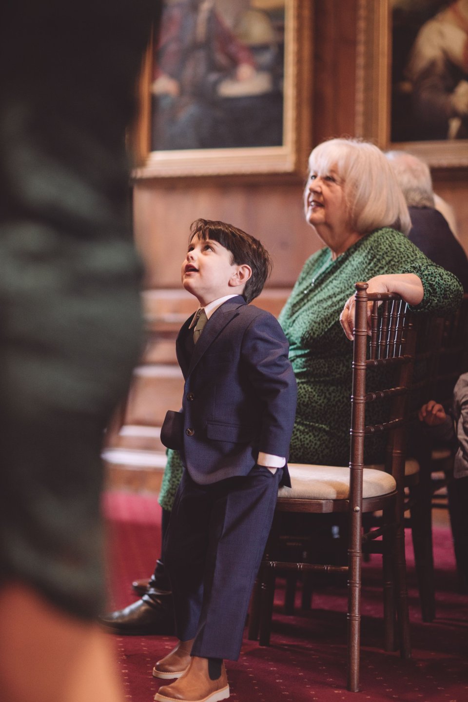 A young boy looks up at his parents as he leans back on a wooden chair with his grandmother in the background.