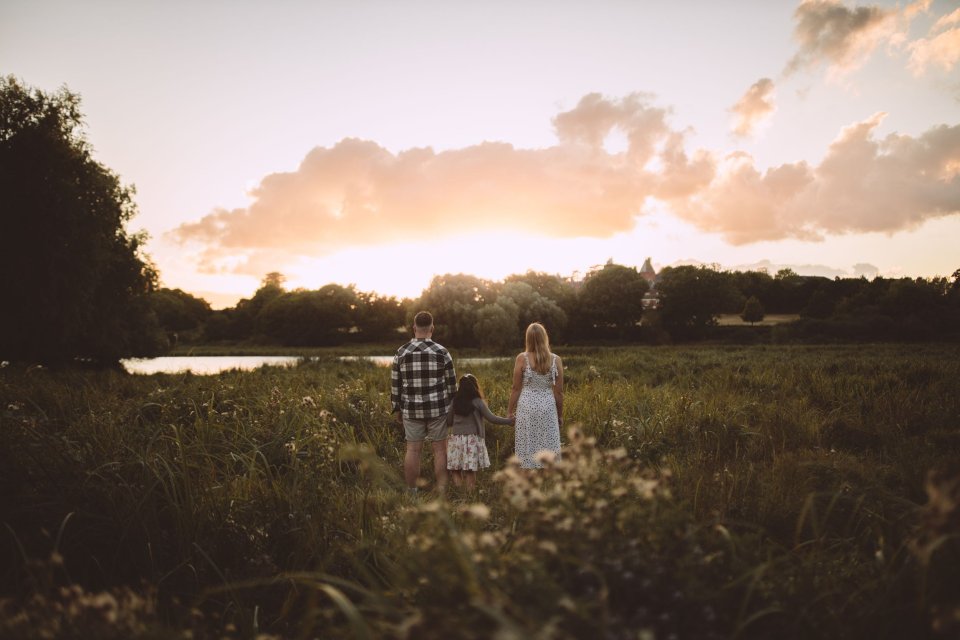 A young man and woman stand side by side in a field of long grass watching the sun set over a lake.