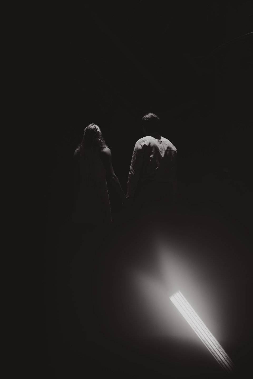 Two silhouetted figures standing in dramatic dark lighting with a bright beam below.
