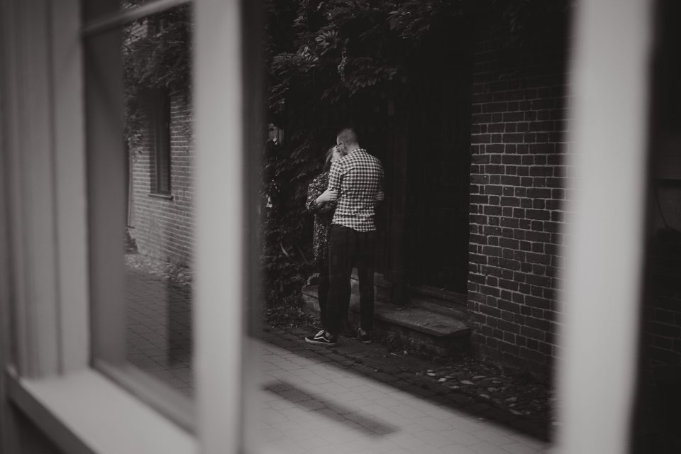 Black and white view through a window showing a figure walking near a brick building entrance.