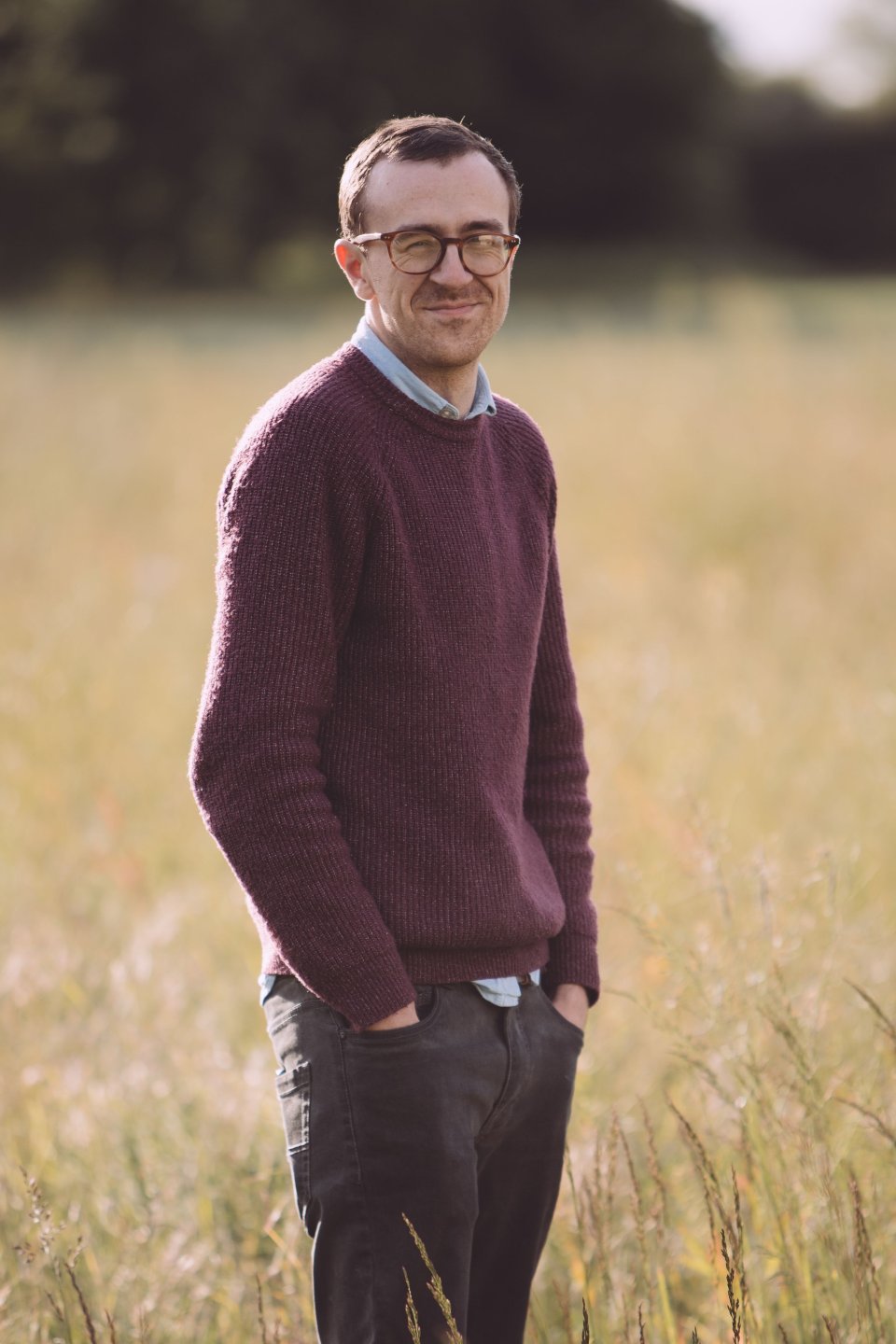 Person in burgundy sweater and glasses standing in tall grass field.