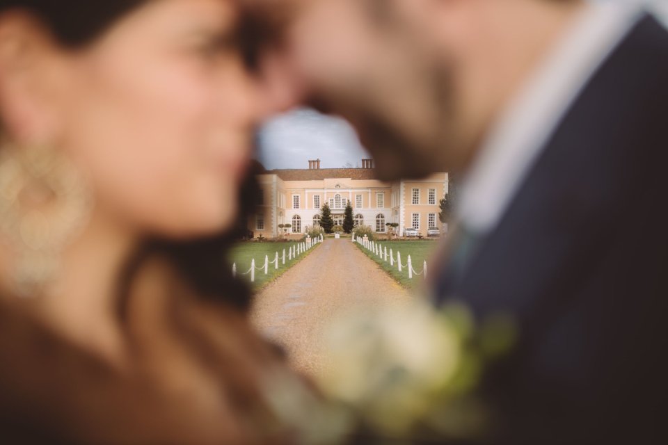 Hintlesham hall is visible through the gap between an out of focus bride and groom who have their heads touching