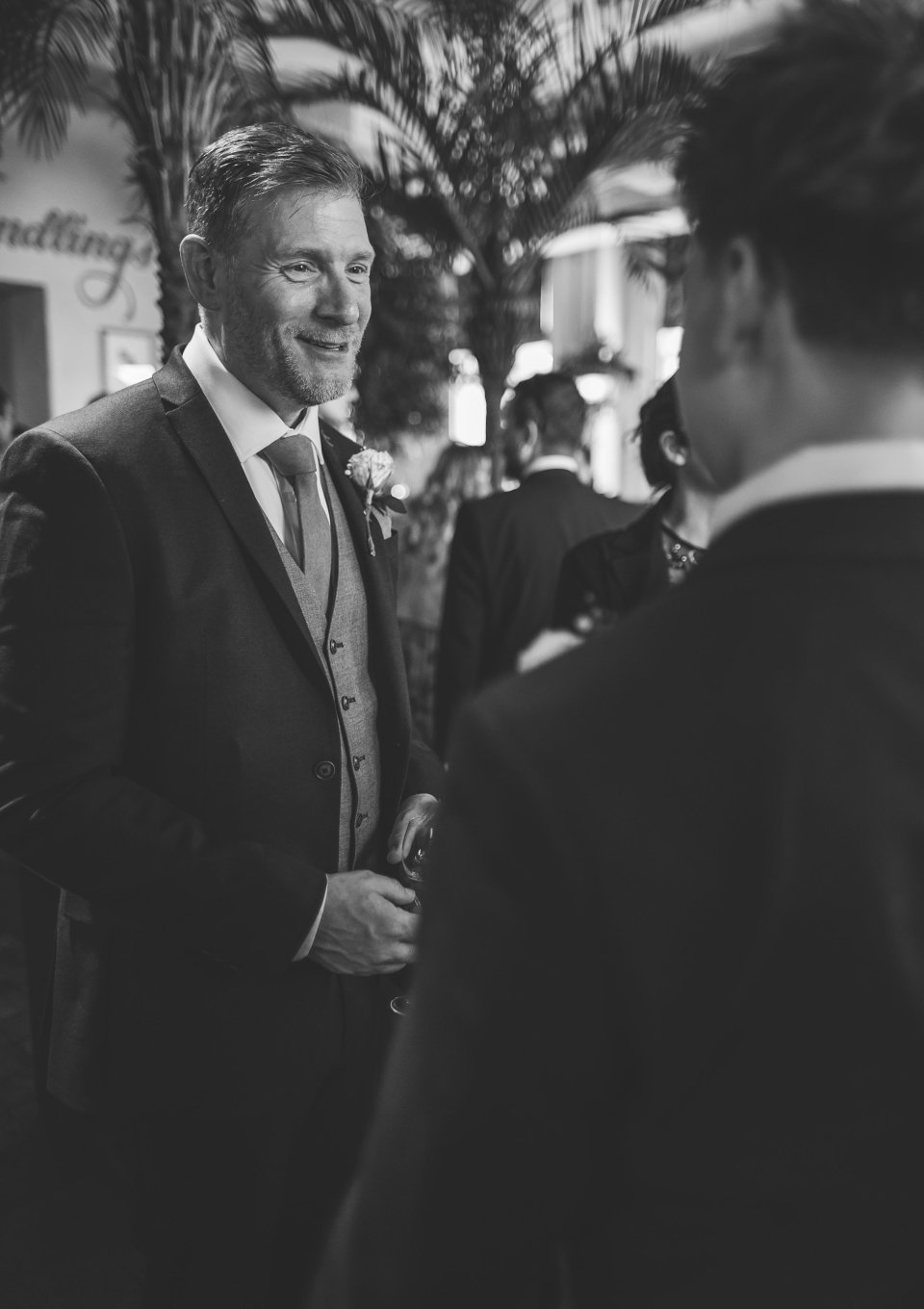 A well-dressed groom in a suit and tie engages in conversation at an elegant indoor reception drinks.
