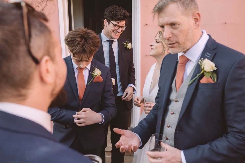 Groomsmen in navy suits with coral boutonnieres stand together against a pink wall during the wedding reception.
