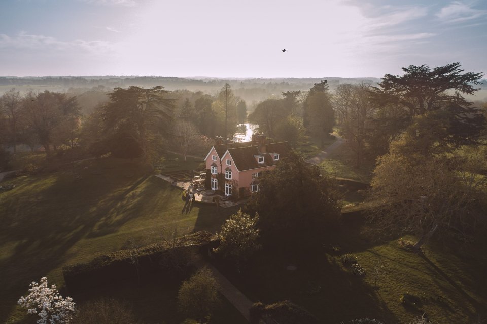 Aerial view of a charming Nedging Hall surrounded by lush green trees on a sunlit hilltop.
