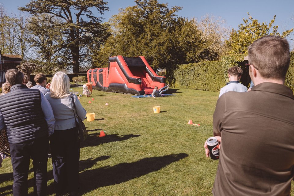 People gather on a lawn watching a large red inflatable bounce house during an outdoor event on a sunny day.