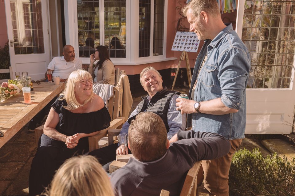 Friends enjoy an outdoor gathering around a wooden table on a sunny patio during daytime.