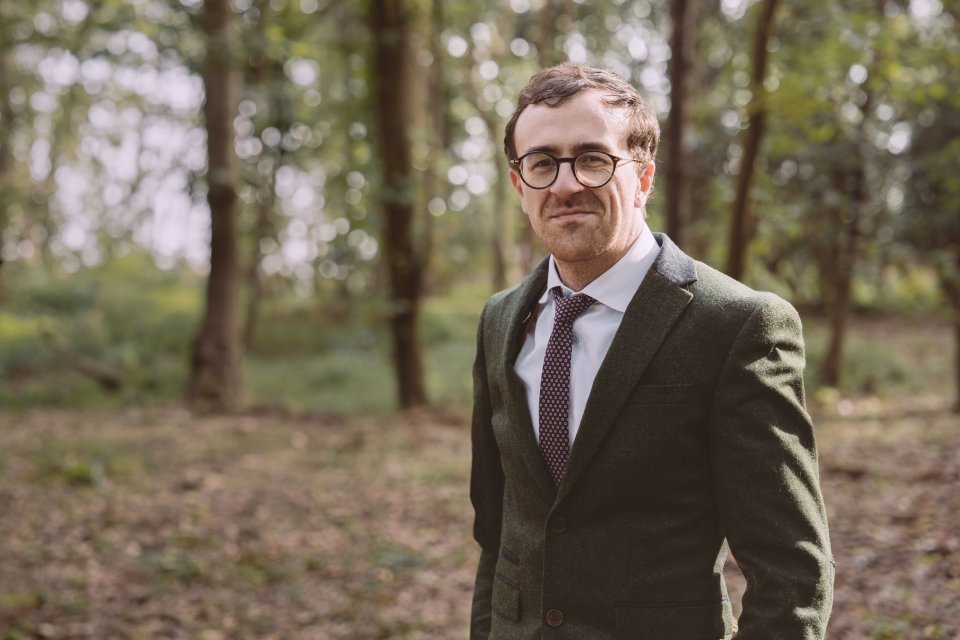 Professional portrait of a gentleman in a brown suit and glasses standing in a wooded outdoor setting.