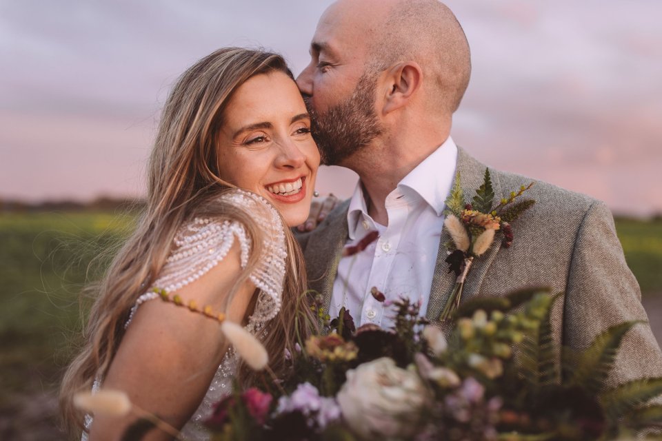 A joyful Suffolk wedding couple embraces in a field at sunset wearing lace and tweed with a floral bouquet.