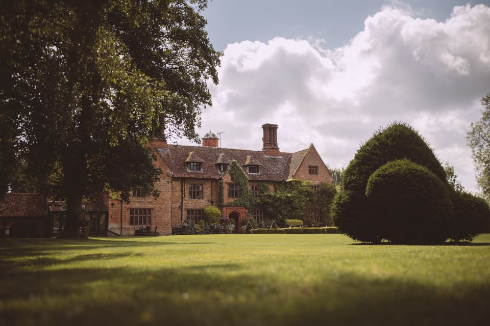 A grand English manor house (Woodhall Manor, Suffolk) with stone facades and manicured gardens stands beneath puffy clouds on a sunny day.