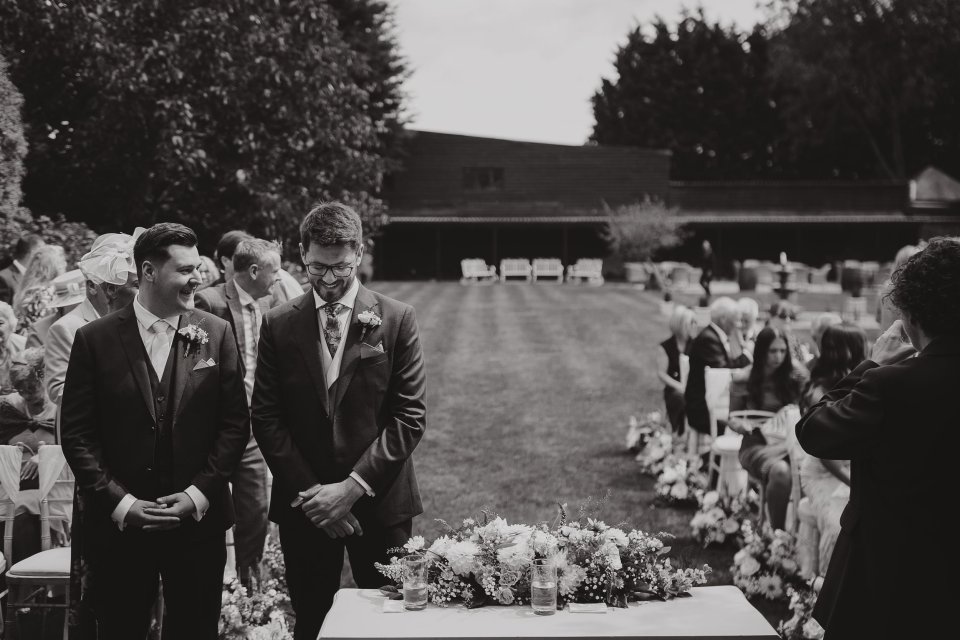 Outdoor wedding ceremony with floral arrangements and guests seated on lawn in black and white.