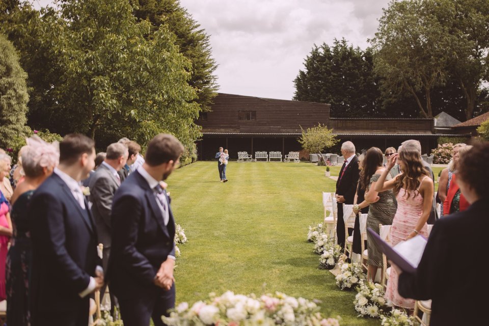 Outdoor wedding ceremony with a flower girl being carried towards the aisle and the congregation turned to watch