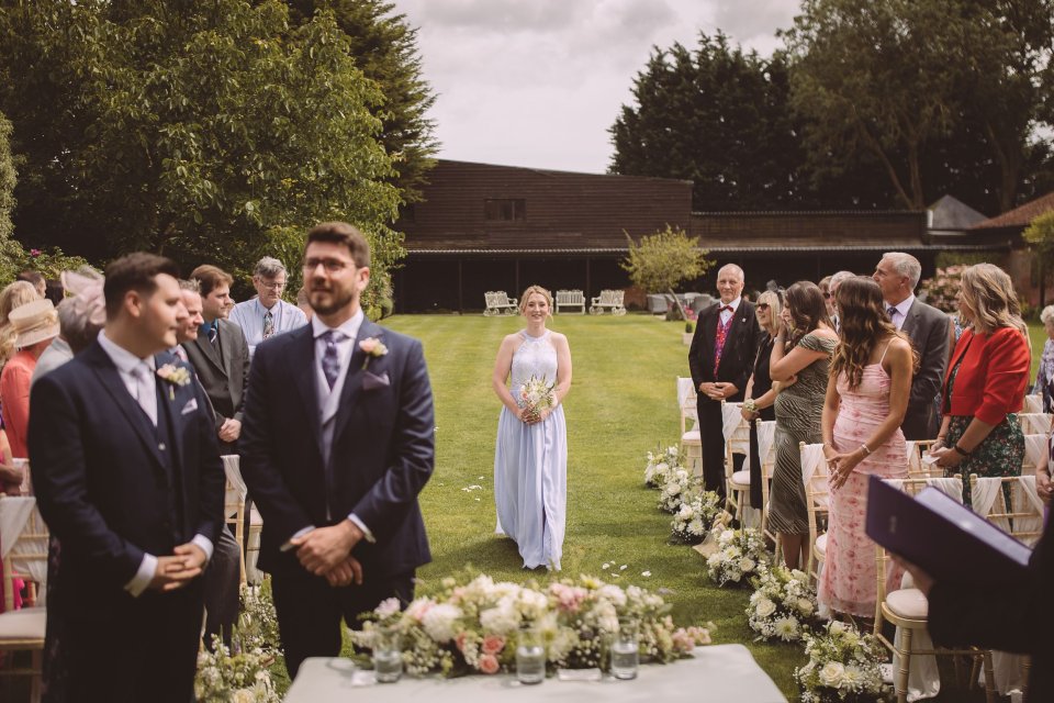 Bridesmaid walks down aisle at an outdoor wedding ceremony with groom in the foreground