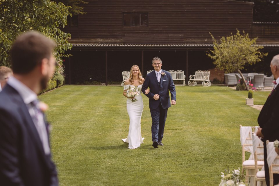 A couple walks down a grassy aisle during an outdoor wedding ceremony with a rustic barn backdrop.