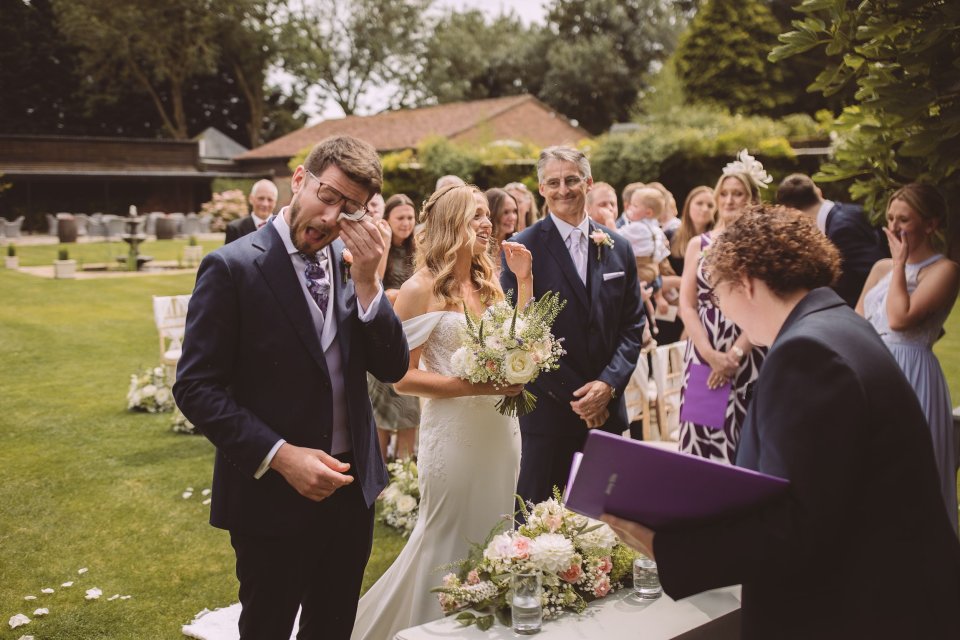 Wedding guests gather on a lawn during an outdoor ceremony near a rustic building.