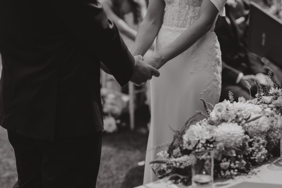 Close-up black and white shot of couple's hands during wedding ceremony with floral arrangements.
