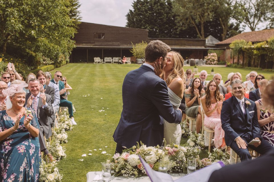 First kiss at a wedding ceremony with guests seated outdoors and floral arrangements in foreground.