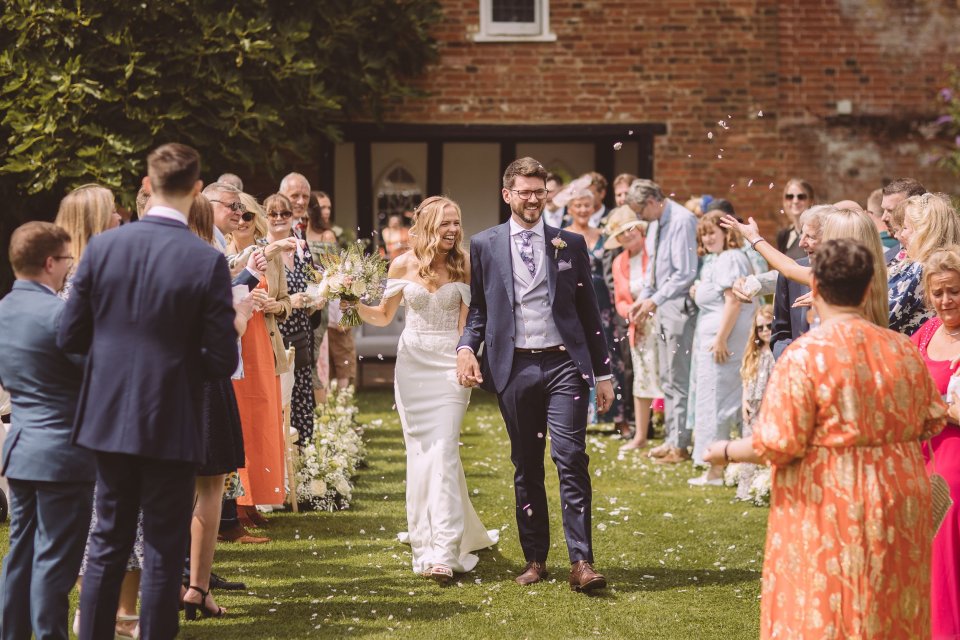 Newlyweds walk down aisle between rows of guests clapping and throwing confetti at a Suffolk outdoor wedding.