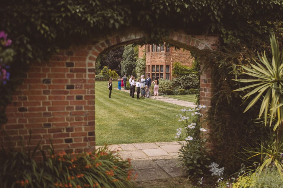 Garden path through a brick archway leading to wedding guests gathering on a manicured lawn.