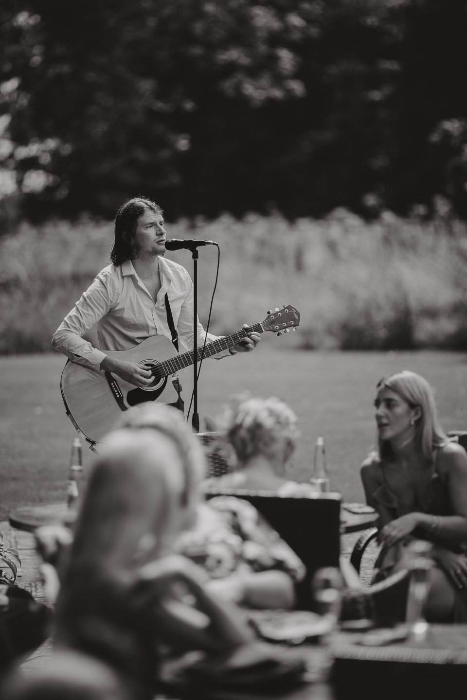 A musician performs with an acoustic guitar outdoors while wedding guests relax.