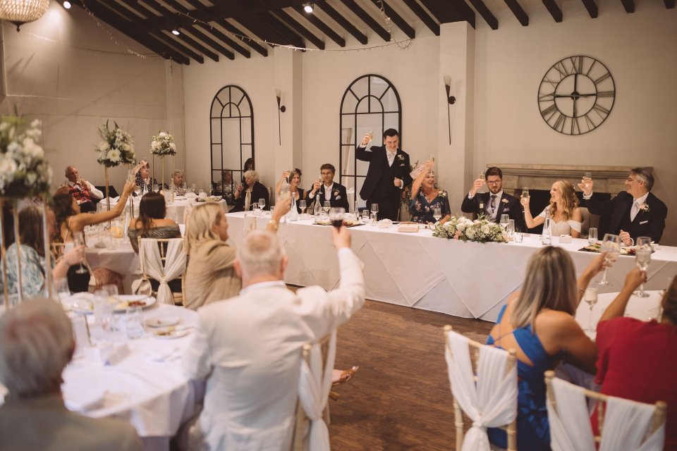 Wedding reception guests seated at long tables in an elegant room with arched windows and a large clock on the wall.