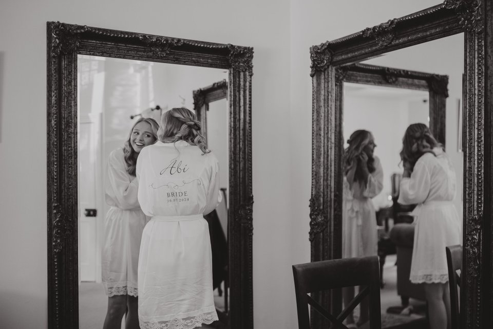 A bride wearing a dressing gown puts on earrings facing a mirror with a second mirror to her right showing a different angle