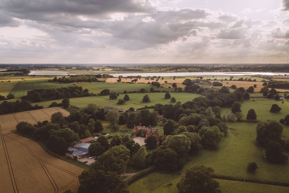 Aerial view of rural countryside with farmland, scattered trees and a stately manor under dramatic cloudy skies.