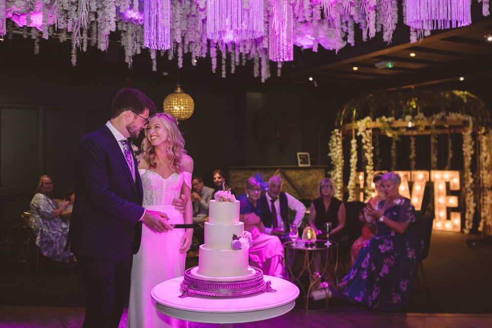 Couple cutting their wedding cake under dramatic purple lighting with hanging decorations.