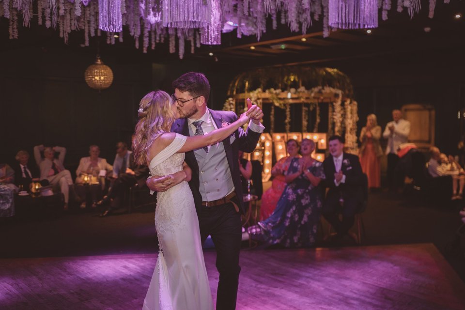 Couple dancing at their wedding reception under purple lighting and hanging decorative elements.