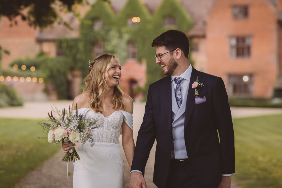 A romantic wedding photo of a bride and groom walking hand in hand with Suffolk Wedding venue Woodhall Manor in the background.
