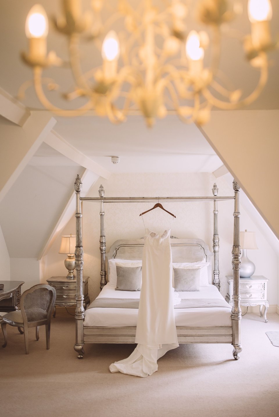 Elegant white wedding dress hangs on four-poster bed in romantic bedroom with crystal chandelier above.