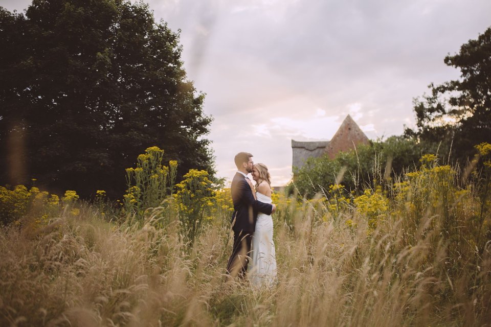 Couple embraces at their wedding in a Suffolk field of yellow wildflowers with an elegant building visible in the distance.