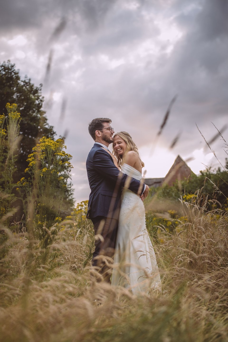 Dramatic wedding portrait of a couple embracing in tall grass with moody storm clouds overhead.