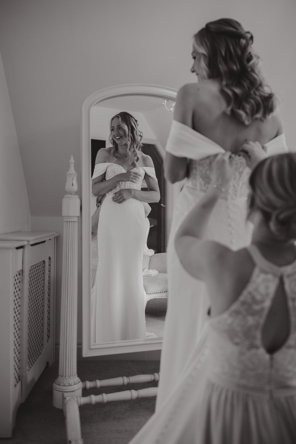 Black and white photo of a bride in an off-shoulder wedding dress looking at her reflection in a standing mirror.