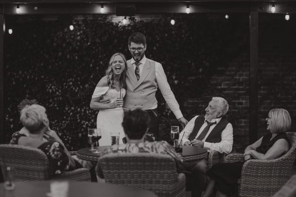 Black and white reception photo showing wedding guests seated at tables with string lights overhead as bride and groom laugh.